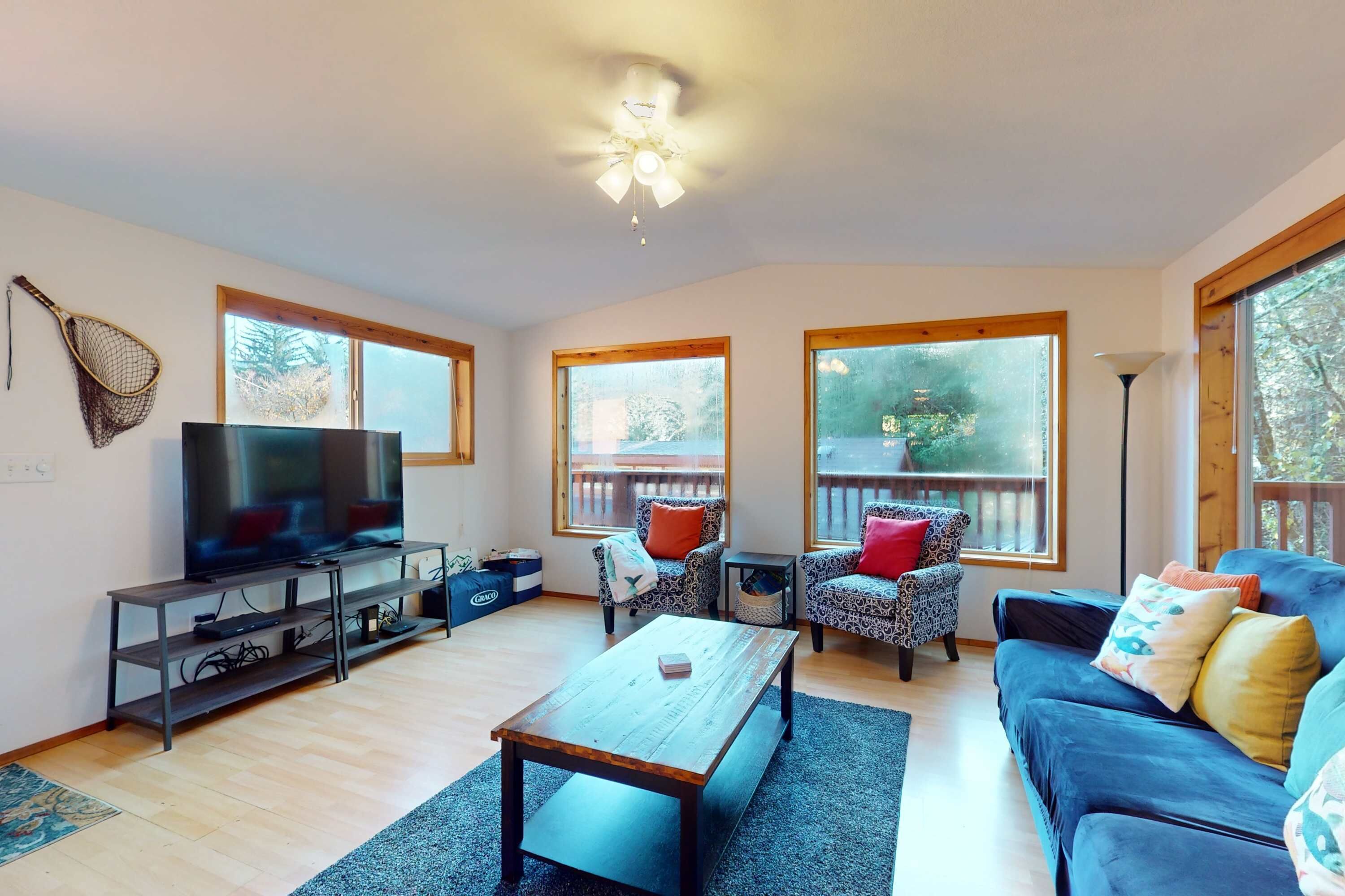 A living room with a TV, sofa, chairs, and large windows.