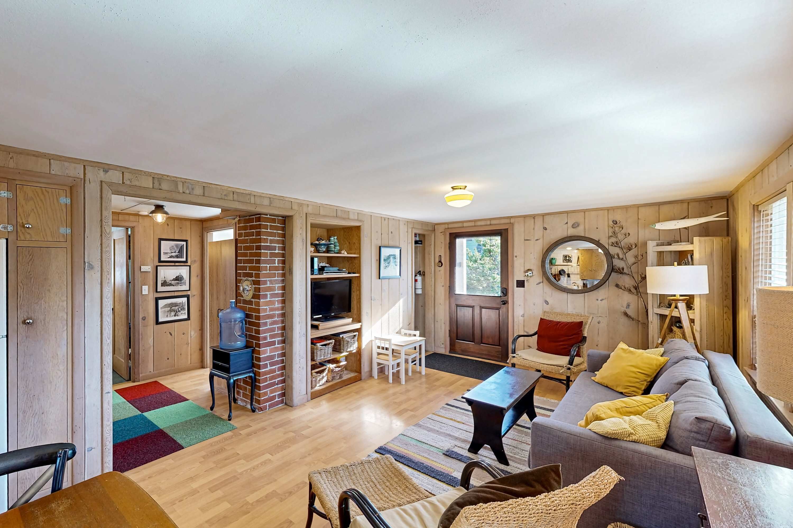 A cozy living room with a sofa, a coffee table, and a TV. There is a brick fireplace, a bookshelf, and a door leading to another room.