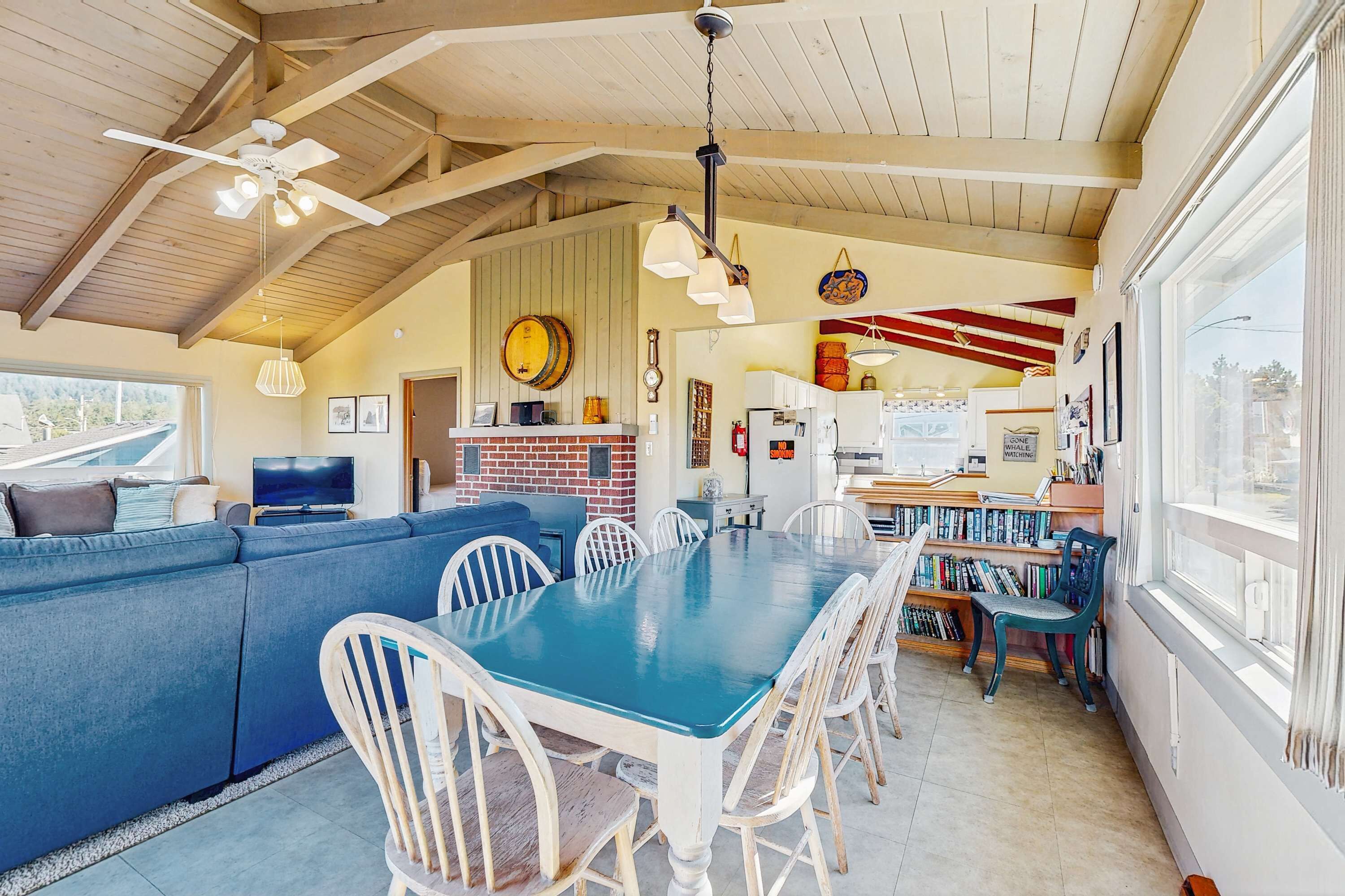 A cozy living area with a blue sofa, a dining table, and white chairs. There is a kitchen area with a brick wall and a window.