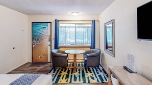 Een kamer met een bed, een klein tafeltje met twee stoelen, een raam met gordijnen, een wandtelevisie en een deur met strandkunst.