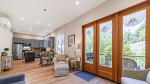 A modern kitchen with a dining area, a sofa, and a balcony with a view of trees. - Newly Built, Dog-Friendly, Colorful Creekside Home - One Mile to the Ocean (Gold Beach)