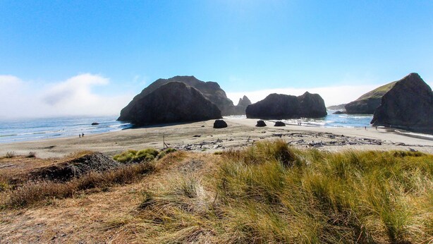 A coastal landscape with large rock formations, a sandy beach, and grassy dunes. - Newly Built, Dog-Friendly, Colorful Creekside Home - One Mile to the Ocean (Gold Beach)