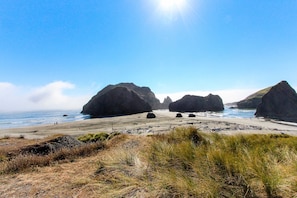 A coastal landscape with large rock formations, a sandy beach, and grassy dunes.