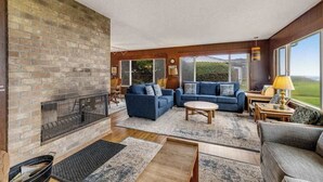 Ein geräumiges Wohnzimmer mit Kamin aus Ziegelsteinen, blauen Sofas und großen Fenstern mit Blick ins Freie.