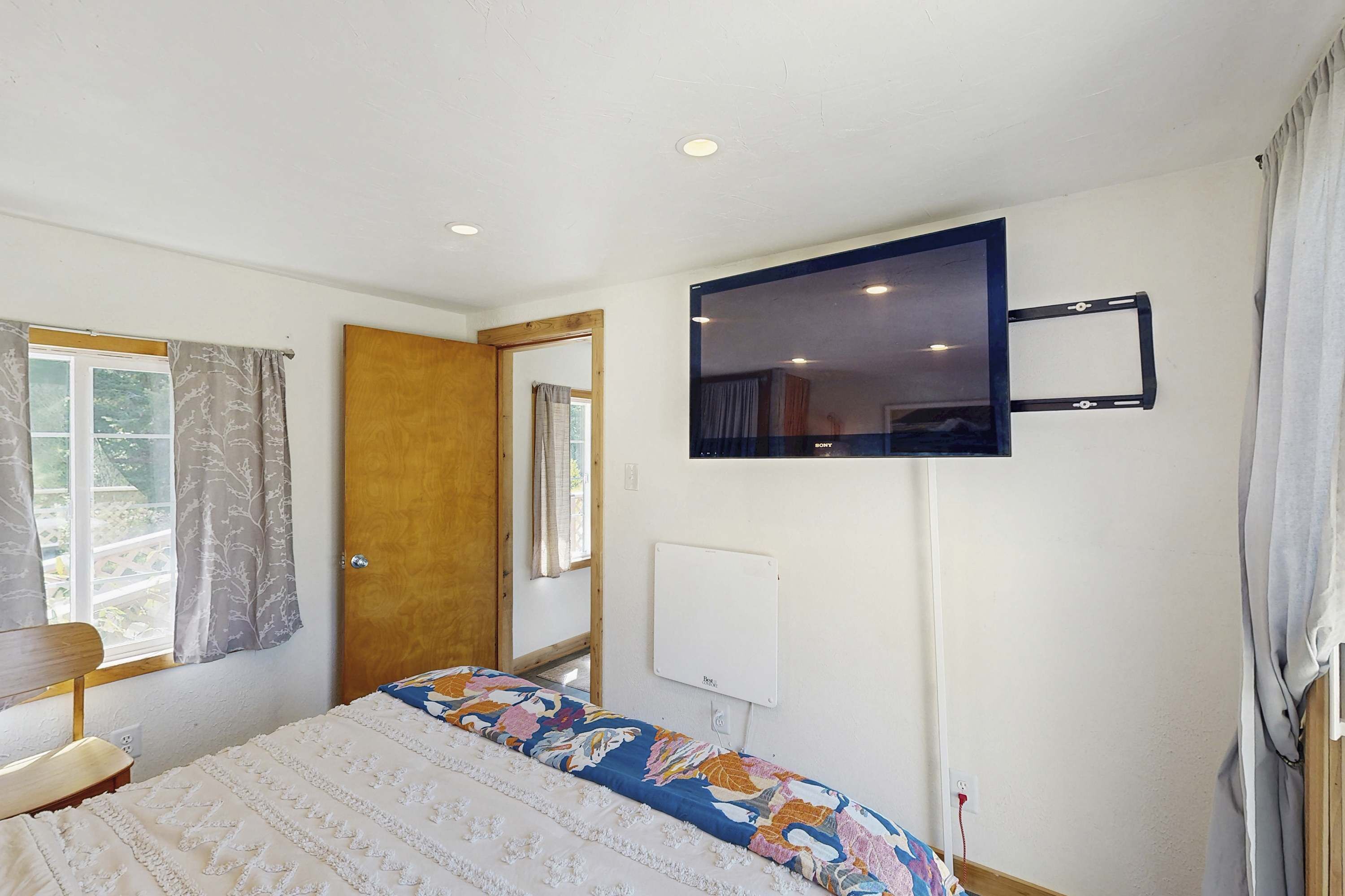 A bedroom with a bed, a television mounted on the wall, a window with curtains, and a door.