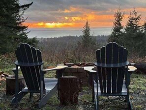 Two Adirondack chairs facing a sunset over a body of water with a forested background.