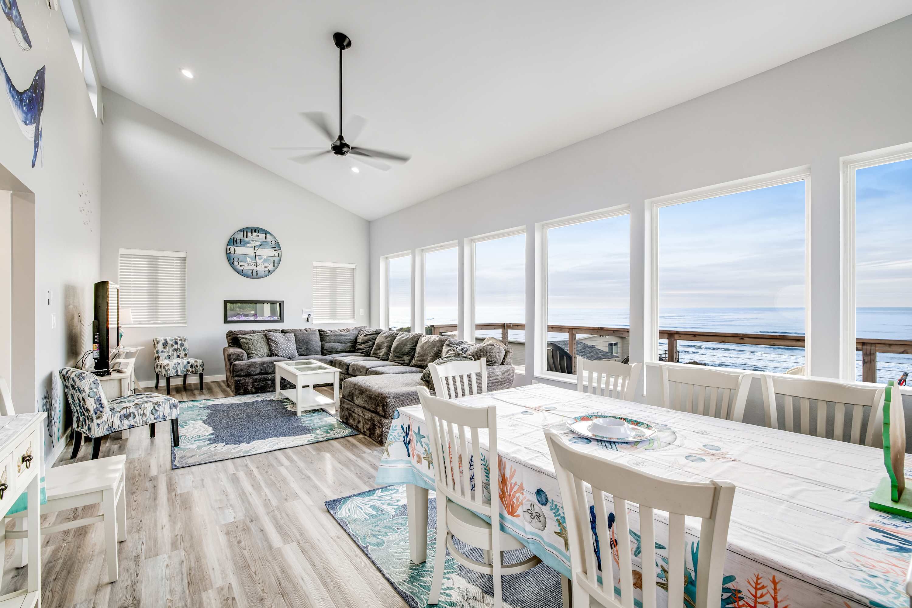 A spacious living area with a large window overlooking the ocean, a ceiling fan, and a dining table set for four.