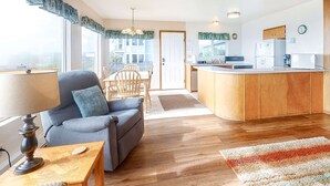 Sala de estar con sofá, una mesa de centro, una mesa de comedor con sillas, una zona de cocina con refrigerador y fregadero, y vistas al exterior.