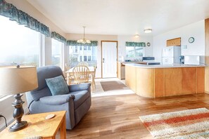A living room with a sofa, a coffee table, a dining table with chairs, a kitchen area with a refrigerator and a sink, and a view of the outside. - Dog-friendly oceanfront home on a bluff with spectacular views (Bandon)