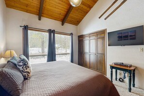 A bedroom with a bed, curtains, a TV, a table, and a wooden ceiling. - Peaceful Cabin Escape | Hot Tub, SHARC passes, A/C, Walk to River - Dogs OK (Sunriver)