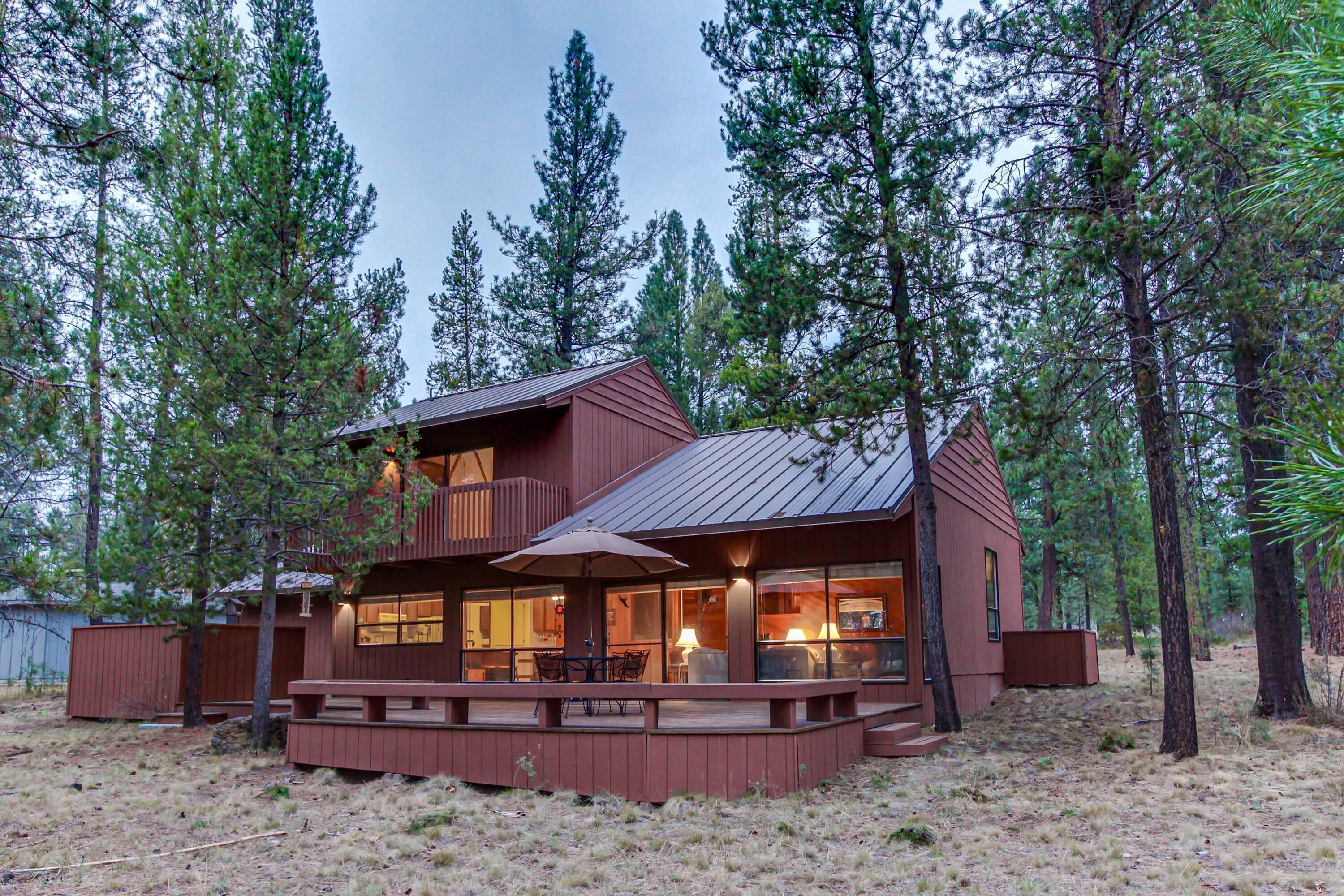 A cabin with a deck, large windows, and a metal roof, surrounded by tall pine trees.