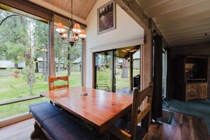 A wooden dining table with benches, a chandelier, and a view of a grassy yard with trees. - Wood-beam cabin with amazing location, pools, hot tub & SHARC passes (Sunriver)