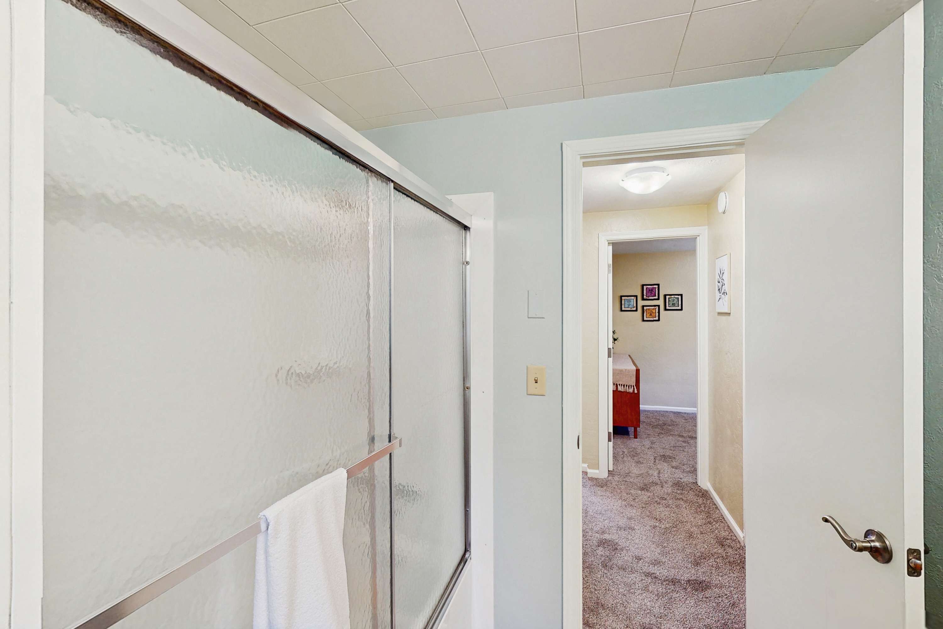 Een hal met een deur van geëtst glas, een witte handdoek aan een metalen stang, en een vloerkleed dat leidt naar een kamer met ingelijste foto’s aan de muur.