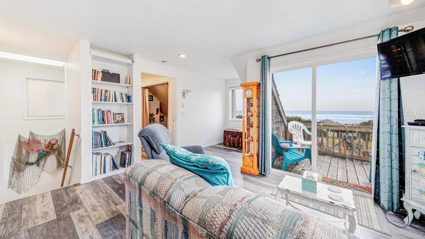 A spacious living room with a large window, a bookshelf, a cozy sofa, and a wooden deck.