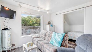 A room with a sofa, a television, a window with a view of trees, a bed, and a small table.