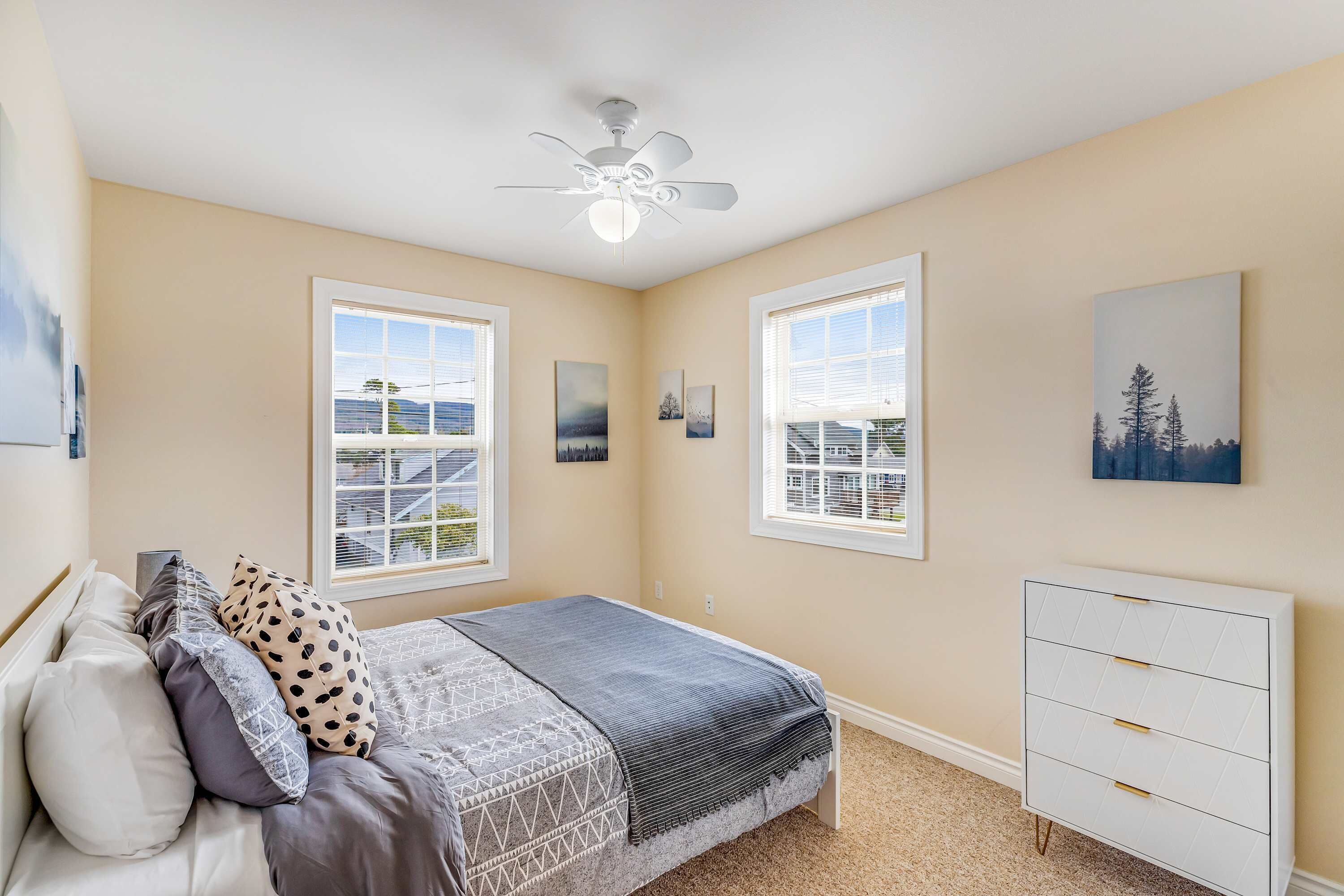 A bedroom with a bed, a ceiling fan, two windows, a dresser, and artwork on the walls.