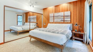 A bedroom with a wooden paneled wall, a large bed, a bedside table, a mirror, and a window with curtains. - Dog-friendly home with grill, deck, firepit, balcony, & mountain view (Black Butte Ranch)
