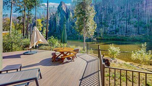 Una terraza de madera con mesas de picnic y una sombrilla, con vistas a un río y montañas boscosas.