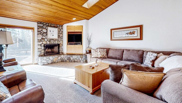 A cozy living room with a stone fireplace, wooden ceiling, and a large sofa. - Spacious home with fireplace, private hot tub, AC, & SHARC passes (Sunriver)