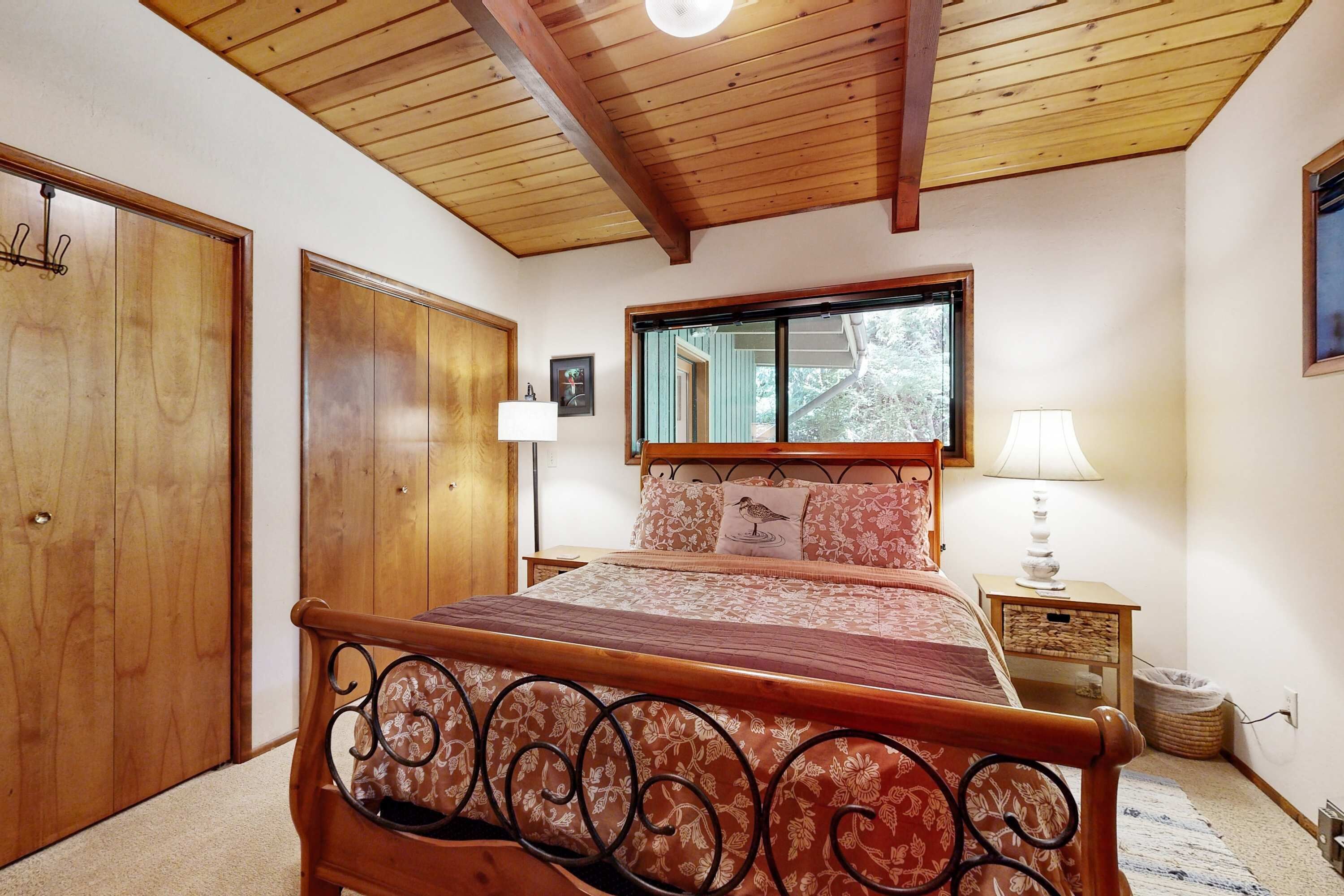 A bedroom with a wooden ceiling, a bed with a metal headboard, a nightstand, and a window with a view of trees.