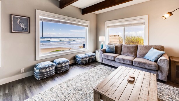 Una sala de estar con un ventanal que da a una playa, un sofá gris y una mesa de centro de madera.