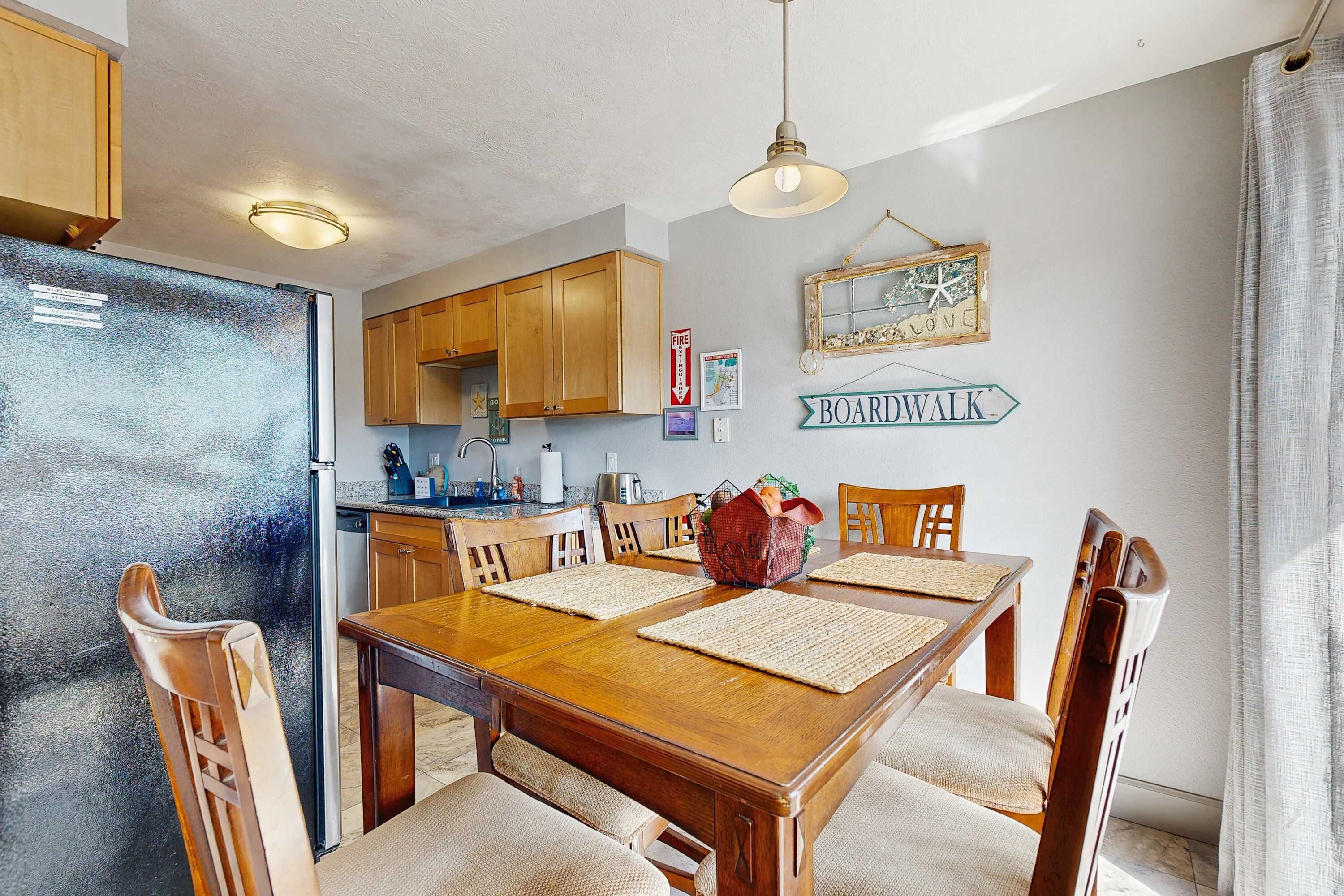 Un comedor con una mesa y sillas de madera, una cocina al fondo y un cartel en la pared.