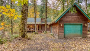 A rustic cabin with a green door, surrounded by trees and a gravel path. - Dog-friendly cabin with deck, fenced yard, game room, & hot tub (Rhododendron)