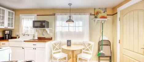 A compact kitchen with a microwave, white cabinets, a dining table with chairs, a broom, and a door.