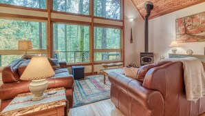 A cozy cabin interior with leather sofas, a fireplace, large windows, and a rustic wooden ceiling.