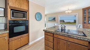A kitchen with wooden cabinets, a microwave, an oven, a granite countertop, a kettle, and a view of the ocean through a window. - Gorgeous dog-friendly home with spectacular ocean views & deck - walk to Main St (Manzanita)