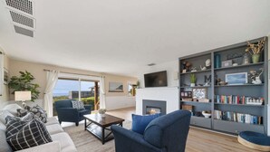 A living room with a sofa, armchair, coffee table, fireplace, bookshelf, and a TV mounted on the wall. - Gorgeous dog-friendly home with spectacular ocean views & deck - walk to Main St (Manzanita)