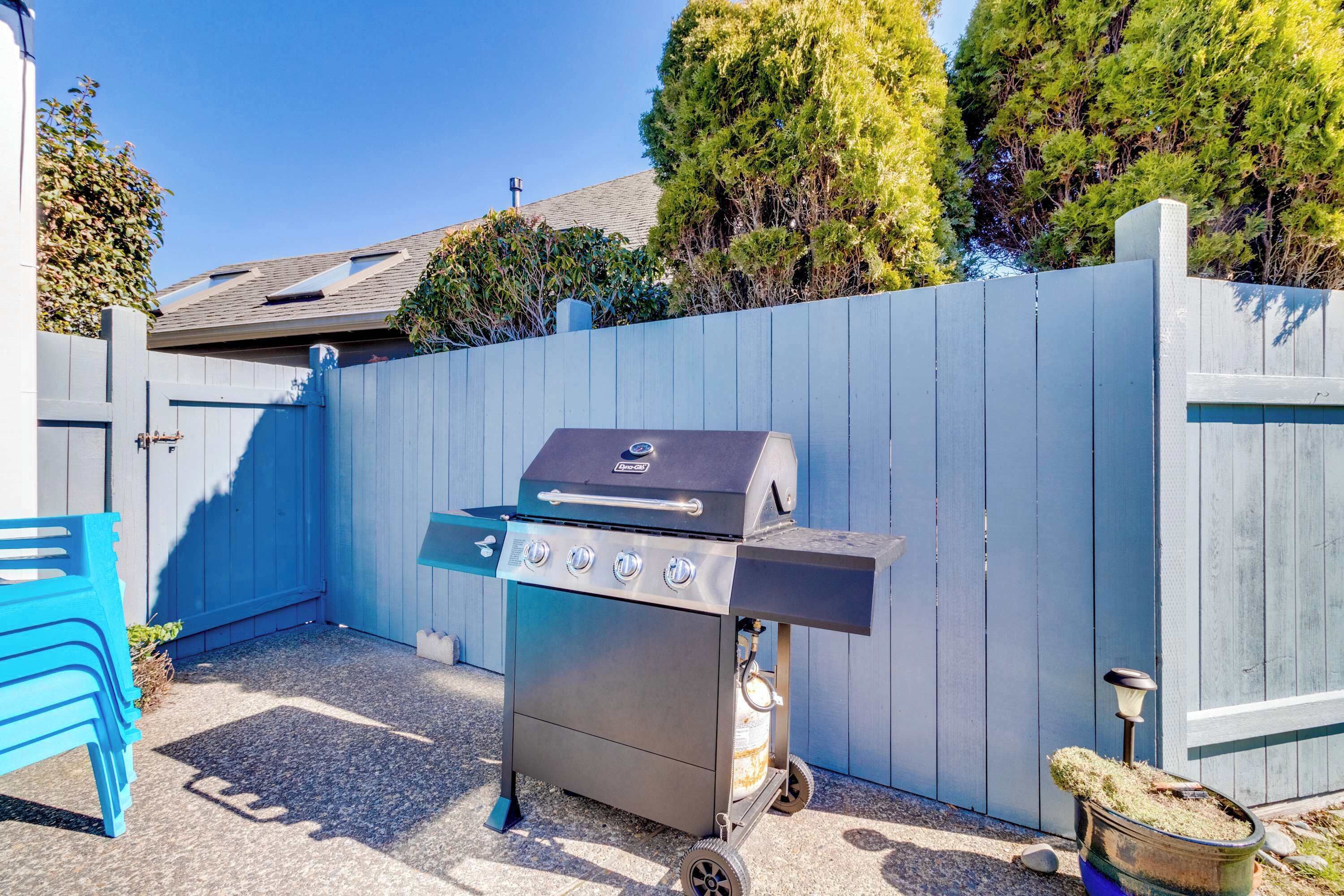 A portable barbecue grill with four burners, a gas tank, and a side burner, positioned in a backyard with a blue fence and a blue chair.