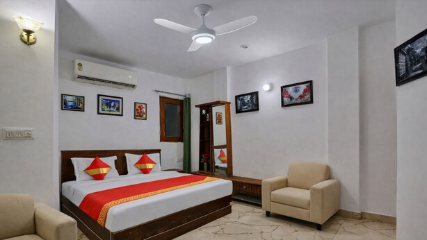 A hotel room with a bed, two armchairs, a ceiling fan, an air conditioning unit, and framed pictures on the walls. - Mauryan Elite I Hotel Hauzkhas  (New Delhi)
