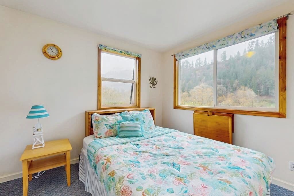 Un dormitorio con cama, una mesita de noche con lámpara, dos ventanas y un reloj de pared.