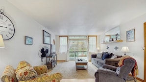 A living room with a sofa, armchair, and a coffee table. There is a large clock on the wall and a window with curtains.