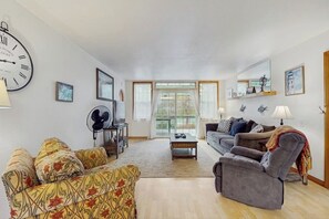 Ein Wohnzimmer mit Sofa, Sessel und Couchtisch. Eine große Uhr hängt an der Wand und ein Fenster mit Vorhängen ist zu sehen.