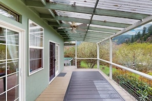 A covered porch with a ceiling fan, large windows, and a view of a garden. - Charming home on the Siuslaw River with a fire pit, WiFi,  & W&D (Mapleton)