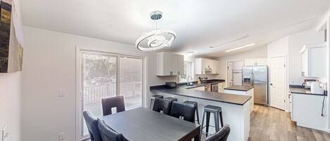 Uma cozinha moderna com ilha central, sala de jantar com mesa e cadeiras de madeira escura, e uma grande janela que oferece luz natural.