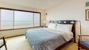 Een slaapkamer met een groot raam, een bed met een beddekleed met patroon, een nachtkastje, een lamp en een stoel.