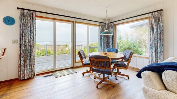 Een kamer met een houten vloer, een ronde tafel met stoelen, een bank en een groot raam met gordijnen.