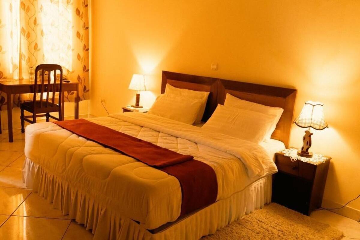 Una cama bien hecha con cabecera de madera, dos lámparas de noche y una silla en una habitación cálidamente iluminada.
