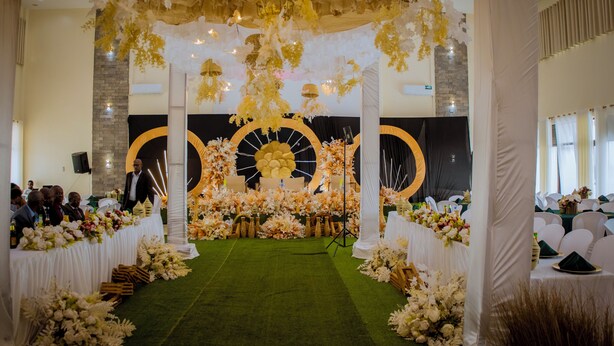 Un salón interior para eventos, con un escenario, arreglos florales y una alfombra verde.