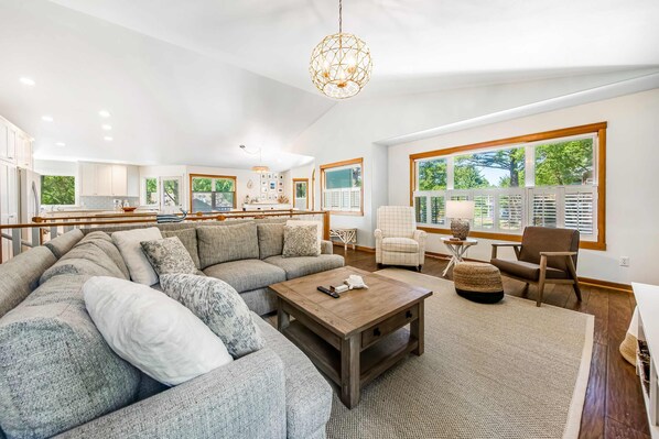 A spacious living room with a large sectional sofa, a wooden coffee table, and a view of the kitchen through large windows. - Spacious home with fenced yard, deck, grill, fast WiFi, & game room (Cannon Beach)