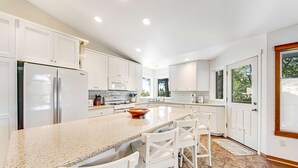 A modern kitchen with white cabinetry, a granite countertop, and a central island with bar stools. - Spacious home with fenced yard, deck, grill, fast WiFi, & game room (Cannon Beach)