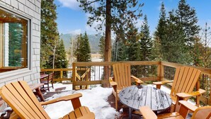 A wooden deck with Adirondack chairs and a table, surrounded by tall trees and a mountain view.