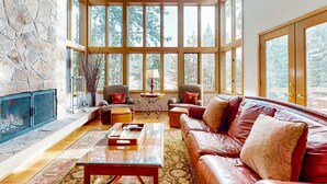 A spacious living room with a stone fireplace, leather sofa, and large windows offering a view of trees.