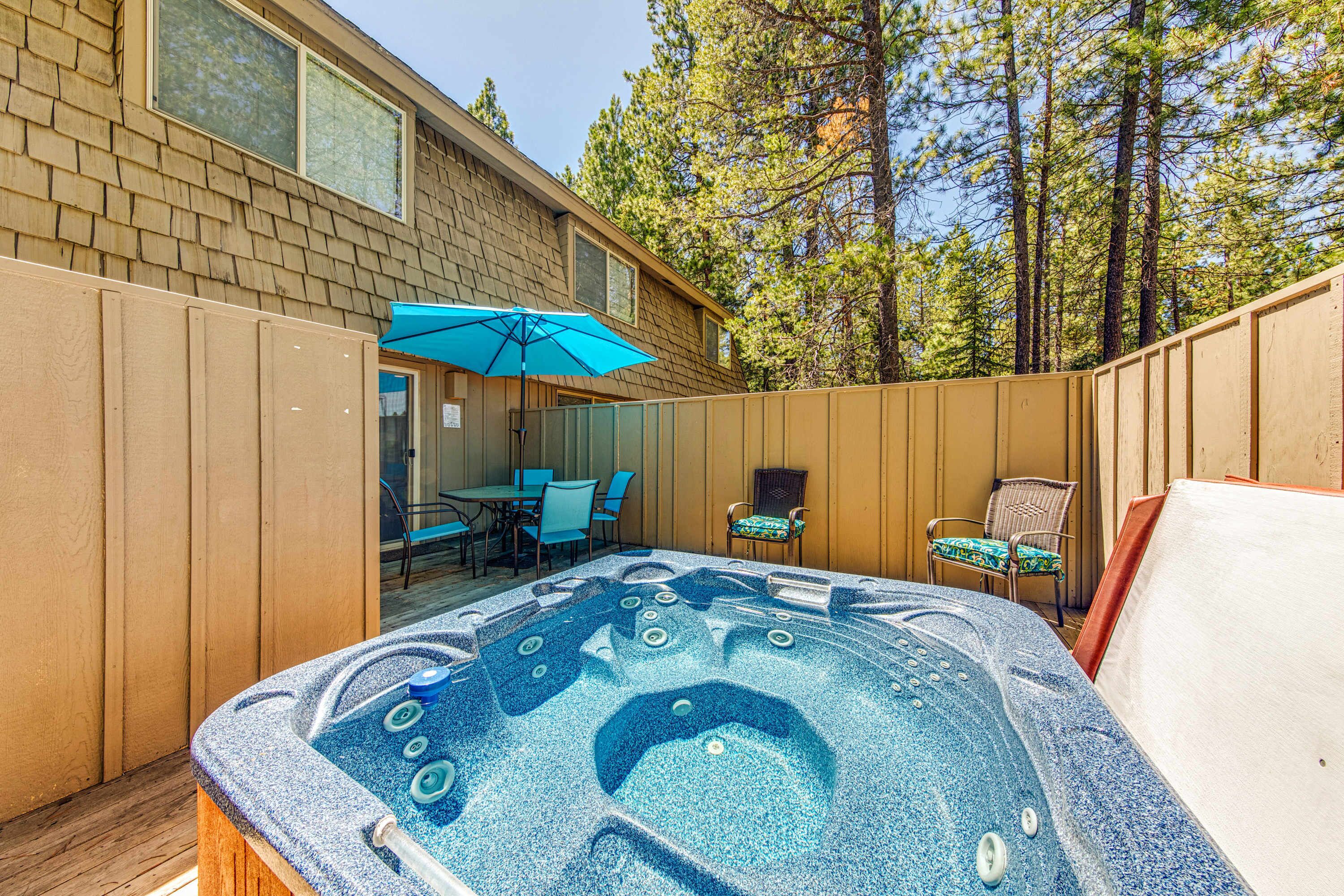 A hot tub in a backyard with a wooden fence, a table with chairs, and a patio umbrella.