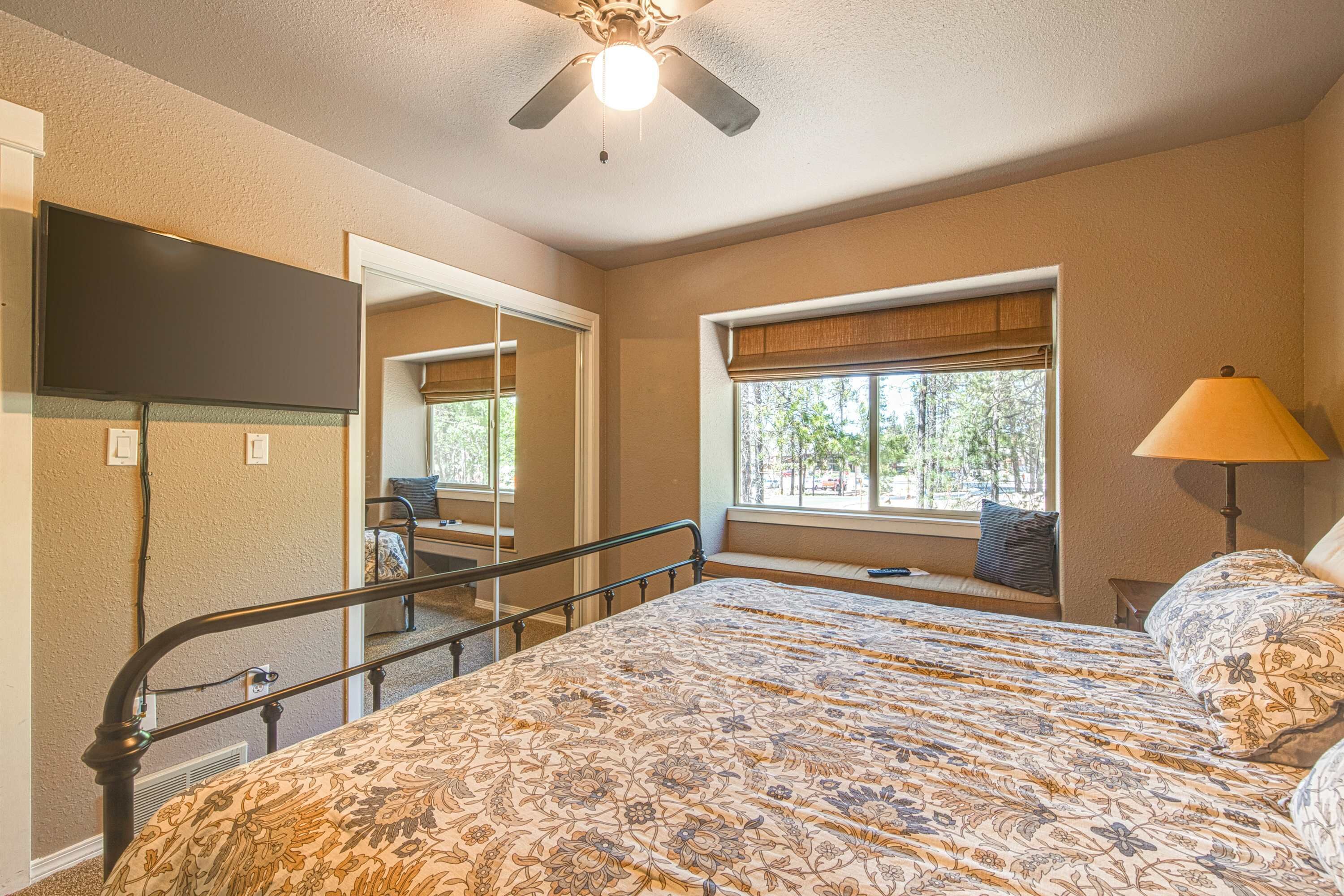 A bedroom with a bed, a ceiling fan, a window with blinds, a lamp, and a television on the wall.