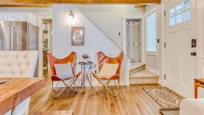 Un interior moderno con techo de madera, una zona de comedor con mesa y sillas, una cocina con refrigerador de acero inoxidable, y una escalera que lleva a otro nivel.