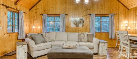 A log cabin interior with a sectional sofa, ottoman, dining table, and chairs.
